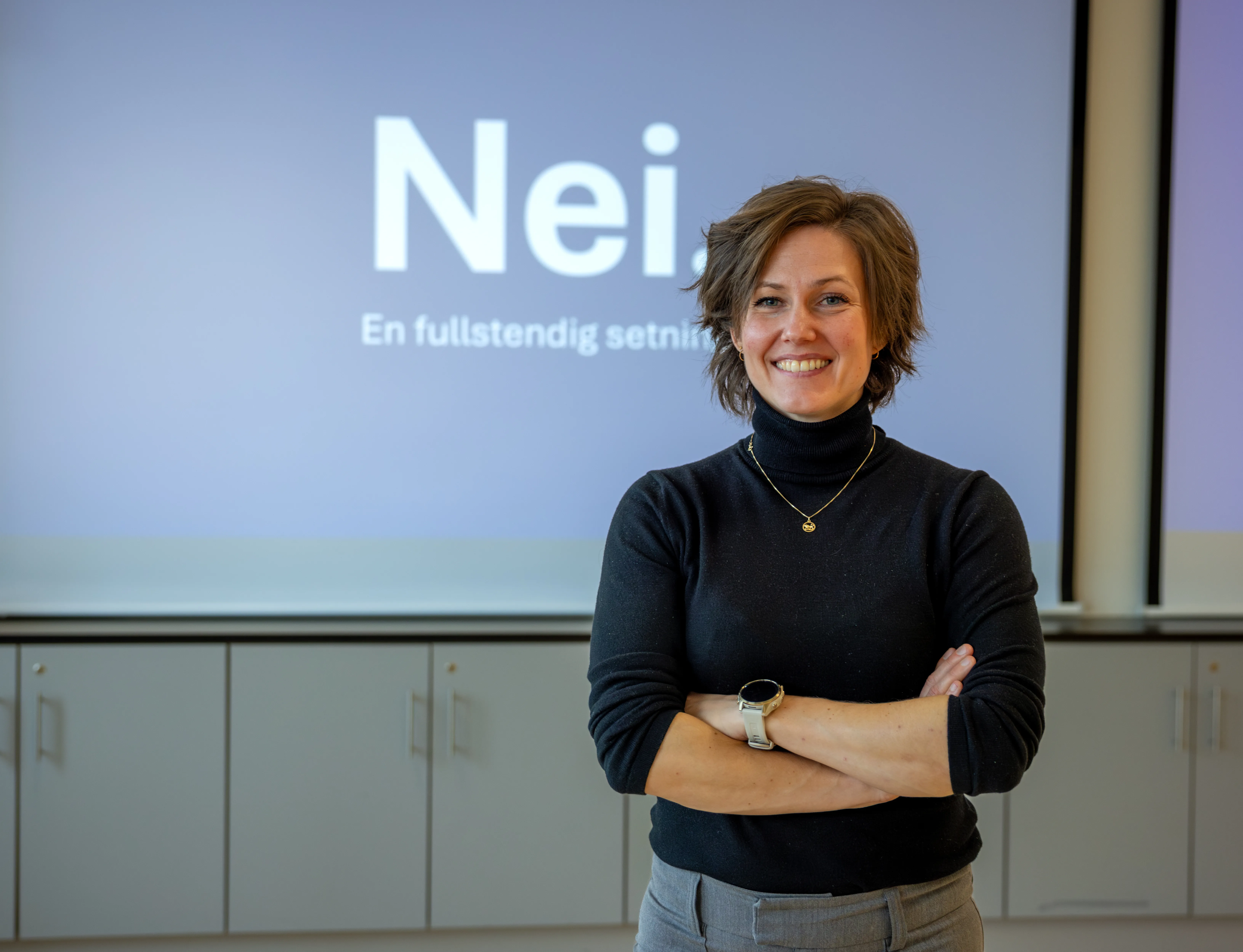 Maya Krogstad står smilende med armene i kors foran et lerret med teksten «Nei. En fullstendig setning».